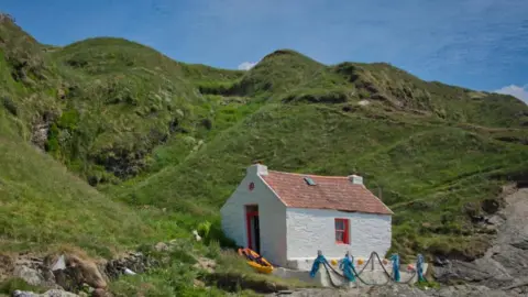 MANX SCENES A small white cottage with red windows and door and an orange roof. There are hills behind it.