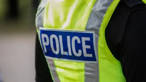 The back of a police officer wearing a high visibility vest with a black jacket beneath it. The the centre of the high vis is a blue rectangle with the word Police written in the centre in white. 