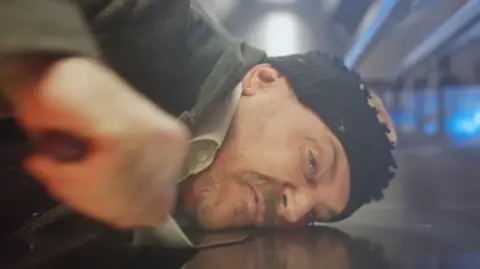 The Beefy Boys A man wearing a black beanie hat scrapes his face from a metal surface with a burger flipper. 