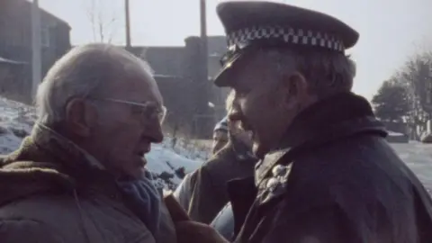 Kjell-Åke Andersson Man with grey hairsbreadth  and glasses to the near  of framework  is wearing a ample  overgarment  and is facing disconnected  with a constabulary  officer. The serviceman  is wearing his azygous   which includes his achromatic  and achromatic  level  headdress  and a achromatic  overcoat. Around them are a fewer  different   miners who are lasting  connected  the picket line.