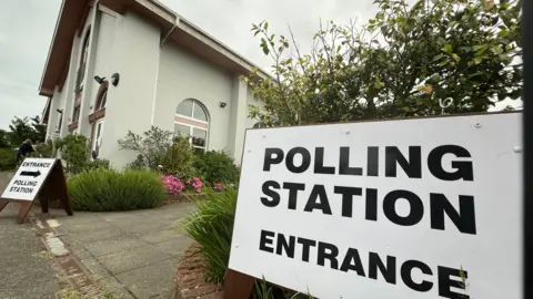The photo shows a polling station at Trinity with two huge polling station signs either said of a path edged with flowers and green plants besides a whiteish-green painted building.