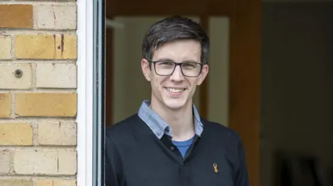 Nick Cook Picture of Nick Cook wearing glasses and a blue top. He is standing in a doorway.