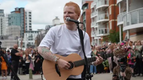 A man with shaved red hair wearing a white T-shirt plays an acoustic guitar and sings into a microphone. There is a crowd of people around him.