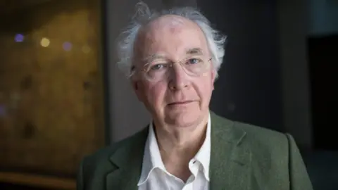 Sir Philip Pullman has wispy white hair and a bald crown. He is wearing a green blazer over a white shirt.