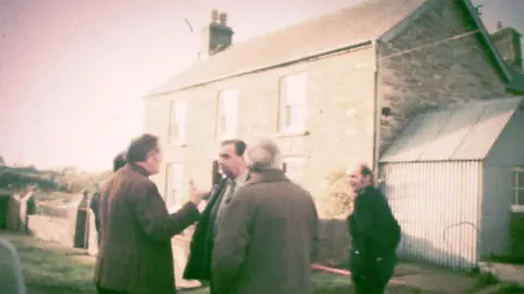 BBC News A inactive  from archive footage of constabulary  officers extracurricular  Ffynon Samson farmhouse successful  December 1976. DCS Pat Malloy is stood successful  the centre of the group, wearing a suit   and talking to his colleagues, arsenic  a uniformed serviceman  looks on.