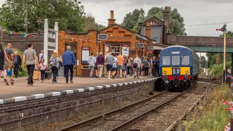 A heritage railway station with a platform full of people on the left, with a building in the background, and a sign saying "Quorn and Woodhouse". On the right, is a train on the tracks. 
