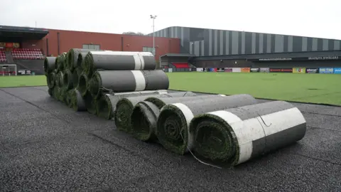 Large rolls of artificial turf stacked on a paved area beside a sports field and stadium seating.