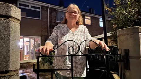 Kevin Shoesmith/BBC Una mujer de unos 70 años, con cabello castaño ralo, posa para la cámara frente a una puerta de metal negra.