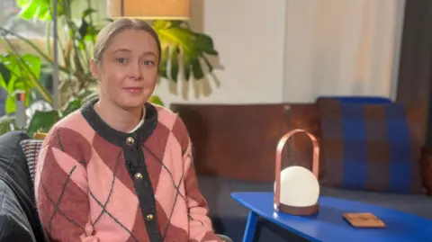 Naomi Campbell wife has on a pink and purple jumper in a diamond pattern and dark trousers. Her hair is tied up. She's looking into the camera ,with a blue table to her left. A lamp is on the table.