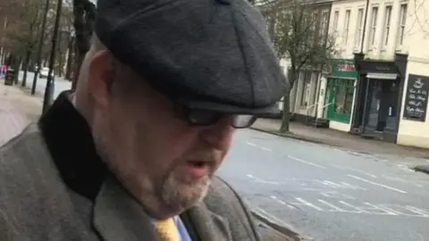 William Kirkbride on his way to court in Carlisle. He is wearing a grey tweed coat, blue shirt and yellow tie. He has a dark grey flat cap on which is pulled over his eyes. He is wearing glasses, has a large double chin and grey stubble. He is walking on a street.