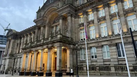 A browny/grey building with five columns at the front which are lit up. there are numerous windows on either side of the columns which go to three floors up. The building is ornate with stone arches above the columns. The sky is grey.