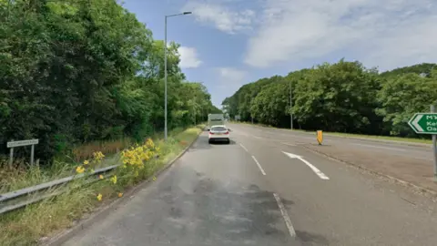 A road as it comes off a roundabout, lined by trees and lamp-posts. There is a sign on the left saying Cottingham Road.