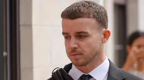 BBC The image shows a person dressed in formal attire, wearing a black suit jacket over a white dress shirt with a black tie. The background appears to be an urban setting with light-coloured stone walls and blurred figures in the distance.