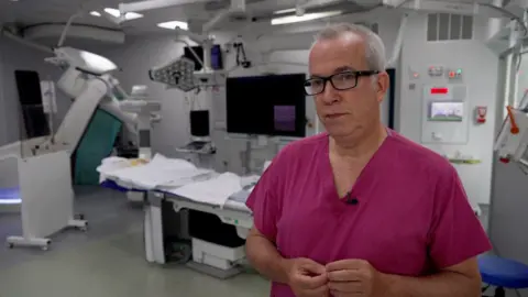 Peter Whittlesea/BBC A surgeon wearing a bright pink shirt and he is standing inside an operation room.