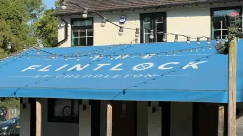 A blue canopy across the front of a two storey cream building. The canopy has the words The Flintlock at Cheddleton written on it