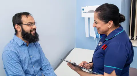 A man with short black hair and beard, wearing a blue shirt, sits on a hospital bed while a nurse with a clipboard talks to him