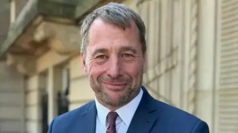 Lancashire County Council An image of Stephen Atkinson. He is standing outside County Hall and is wearing a navy suit, white shirt and red tie. He is smiling.