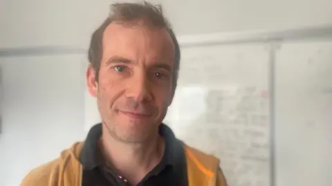 A man with short brown hair wearing a black polo shirt and yellow hoodie standing in front of a white board with writing on it.