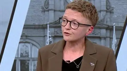 Bev Craig, wearing glasses and a brown jacket over a black top 