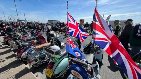 Several motorbikes and scooters parked up in a line. The closest one to the camera has two Union Jack flags at the back as well as a blue RAF flag. There are hundreds of people walking past looking at all of the motorbikes.