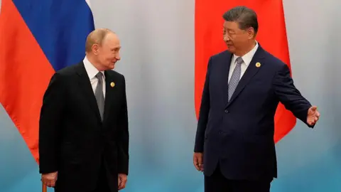 Getty Images Vladimir Putin stands next to Xi Jinping, who olds his left arm outstretched. Behind them are two flags