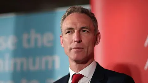 Getty Images Jim Murphy wearing a neutral expression in a dark suit and red tie against red and blue background at a Labour Party event in 2015