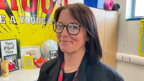 A view of Nicola Evans stood inside the room used to host young carers support sessions. In the background is a wall poster in yellow with PROUD TO BE A YOUNG CARER written on it.