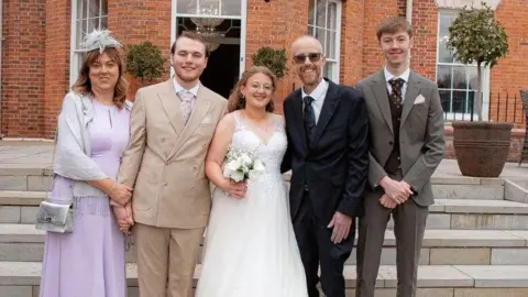Family photo Group picture of Damian Barr with his wife, son, bride and best man at a wedding. They smile in front of a historic venue with trees in plant pots behind.