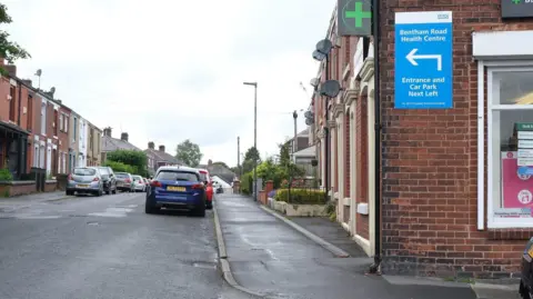 A terraced residential street on Bentham Road with cars parked on either side. There is a pharmacy building in the foreground to the right, with a sign saying 'Bentham Road Health Centre' on the front.