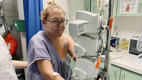 Lindsay Bowers Lindsay Bowers is looking at the camera, wearing dark framed glasses and a hospital gown. She has several wires attached to her and is attached to a few hospital machines.