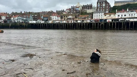 Kadassi Nembhard A woman sat down on a muddy beach, close to the water's edge. She has her back to the camera. On the other side of the harbour are lots of houses.