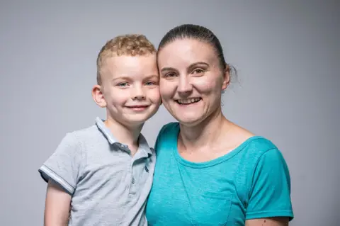 NHSBT A portrait photo of Aiden and mum Steph. He has short blonde hair and wears a grey collared short-sleeved shirt. Steph has dark hair tied back, and a blue t-shirt. They are smiling, their cheeks touching.