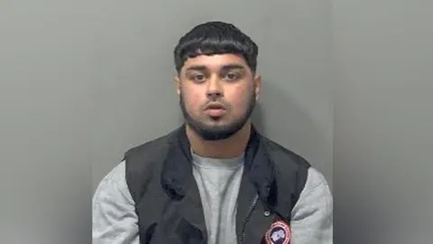 A mugshot of Hamza Arshad, who has a bowl haircut and a smart short beard. He is wearing a pale grey sweater and a black gillet over the top