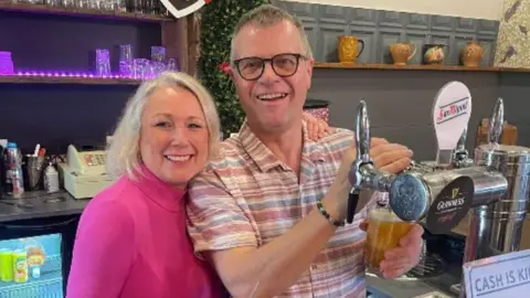Claire in a bright pink top standing next to Neal in a pastel-coloured t-shirt, both behind a bar. Neal is pulling a pint.