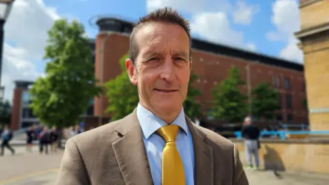 Paul Moseley/BBC Nick Taylor wearing a light brown jacket over a blue shirt, with a golden yellow tie.