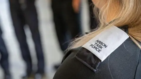 Northamptonshire Fire and Rescue Service The shoulder of a woman who is looking away from the camera. She has blonde hair and the lapel on her jacket says, ARSON TASK FORCE.
