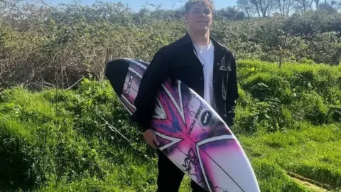 Malakai Hagley The picture shows an individual standing in an outdoor rural setting. The person is holding a surfboard against the body. The surfboard has a bold design made up of pink, white and black sections, with shapes that resemble a stylised Union Jack pattern and some dark graphic markings on the lower part. The individual is wearing a dark jacket over a white top and dark trousers.

