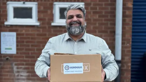 Himmah Sajid Mohammed, director of Himmah, standing outside a building holding a cardboard box.