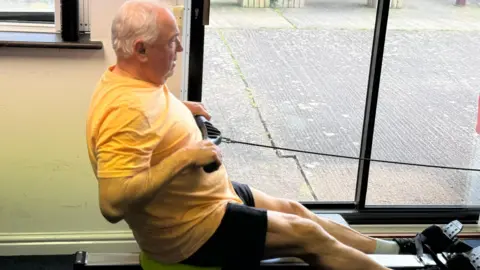 Lester Murphy A man with short grey hair is wearing a yellow T-shirt and black shorts and pulls back as he uses a rowing machine. The rowing machine is positioned next to a patio door which looks out onto a paved concrete area.
