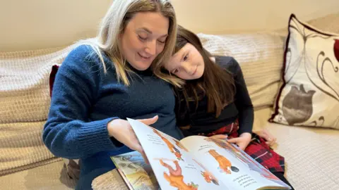 NIKKI FOX/BBC Sarra Wilson reads a book with her daughter Isla, who is resting her head on her mum's shoulder. They are sitting on a sofa.