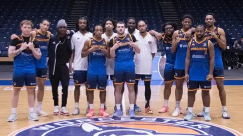 A group of 13 men all dress ing basketball blue and white clothing are stood in a row and smiling at the camera. Some are making peace sign gestures. They are stood on a basketball court.
