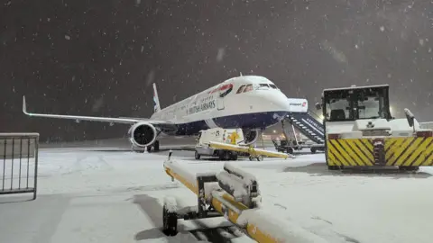 Newcastle Airport A British Airways plane is on the runway as snow falls around it. Snow blankets the ground.