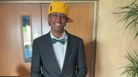 Family photograph Eddie in a mid-shot, smiling to camera, wearing a bow tie, white shirt and black jacket, and a yellow baseball cap side-on