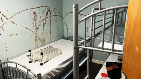 A dorm room of a scout hut with a metal bunk bed and single bed. The green walls are covered in read sauce. Two of the beds have mattresses on them with white sheets, the top bunk does not have a mattress. The single bed has a mobile radiator laid on it. The lower bunk has a black bag and litter on it. The wooden door is slightly ajar.