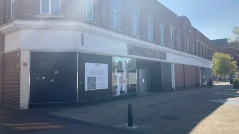 BBC The unused M&S store in Crewe town centre. The outside has boarded over windows in front of a paved area with a bollard in it. A sign on the outside has the M&S logo on it and text underneath. Sunlight is reflecting off windows above the facade.