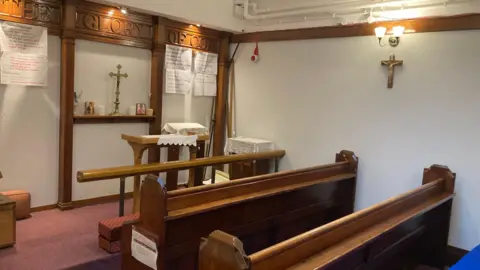 Sally Fairfax / BBC The chapel. The picture shows three wooden benches facing a small altar. The walls are white and there is a golden cross hanging on a wall to the right of the picture.