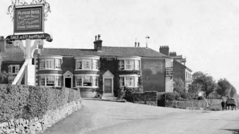 Mitchells and Butlers A black and white photograph of The Plough in Endon. There are three bay windows on the ground and first floors of the building. There is a person walking alongside a horse and cart on the right hand side of the image.