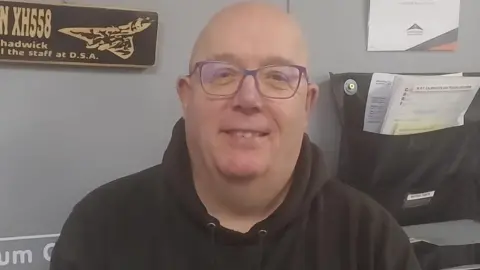 Mark Chadwick looks at the camera. He has a bald head and is wearing glasses and a black hooded top.