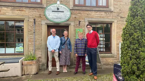 BBC Three members of the Hesket Newmarket Community Shop Project with local MP Markus Campbell-Savours outside the village's old brick shop. Left to right: a bald man with a short, trimmed grey beard and glasses, wearing a light blue button-up shirt over a dark blue T-shirt, light brown trousers and dark brown shoes; a woman with short, light hair, wearing a blue denim jacket over a long, flowing dress, with a bag strap hanging diagonally across her front; a second woman with short, blonde hair, wearing burgundy trousers and a button-up shirt with a purple, yellow, green and blue tartan print; and a very tall man with short, dark hair and glasses, wearing a bright red jumper over a white collared shirt and a dark tie.