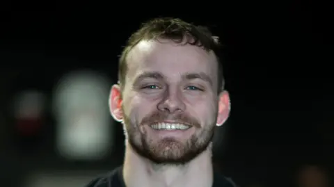 Jamie Niblock/BBC Callum Foster with short dark hair and a beard smiles at the camera. He is wearing a black top and the background is dark and blurred.